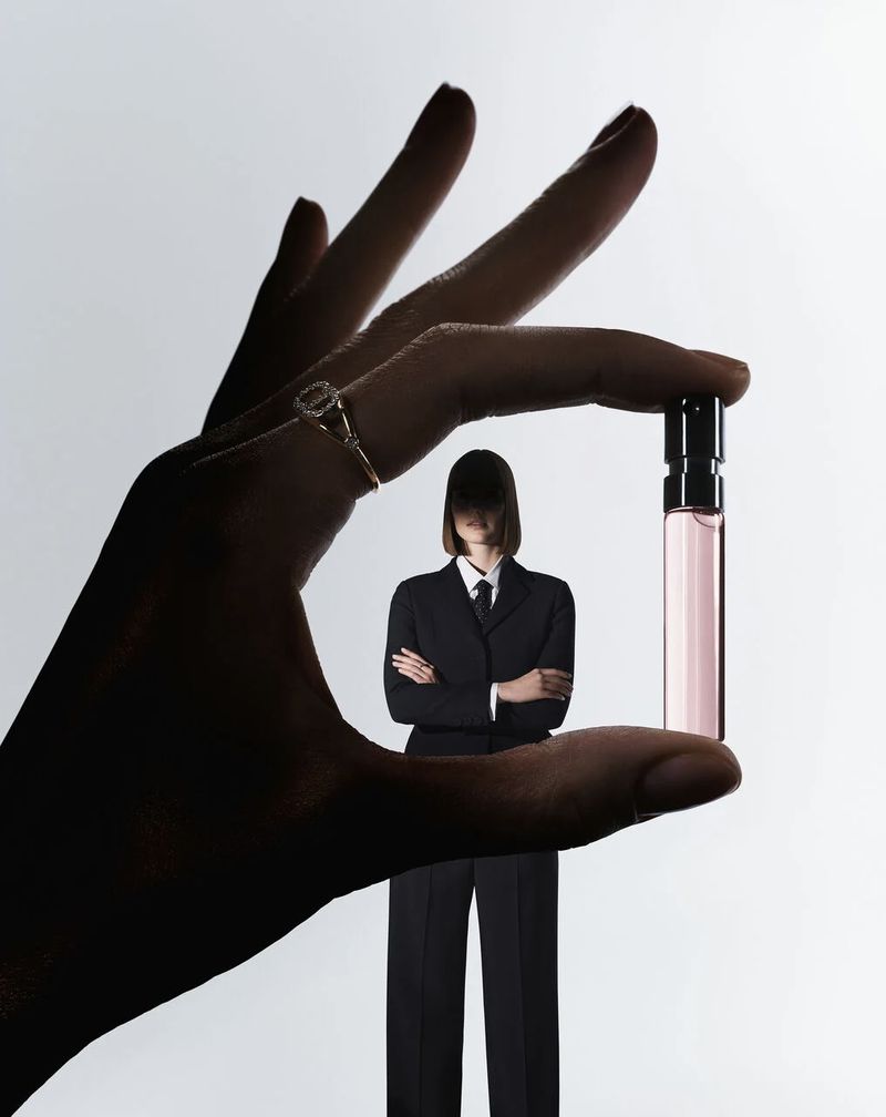 Close-up of a hand holding a small perfume sample vial, framing a person in a suit standing in the background