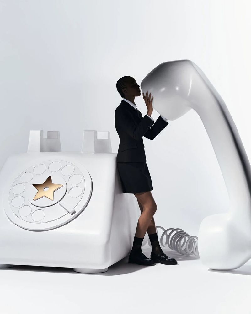 A Black female model in a business suit leans against an oversized, white retro telephone in a white studio setting with clean, modern lines.