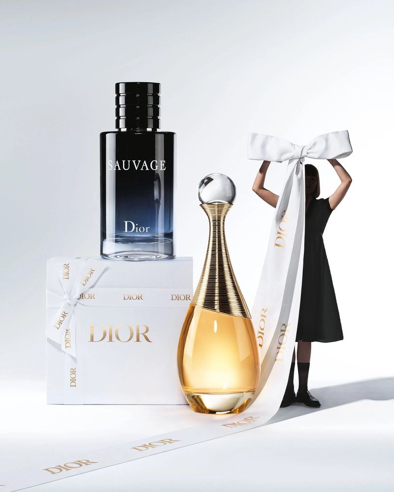 Two Dior perfume bottles are displayed on a white surface, with a person holding a white bow in the background.