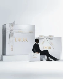 Model seated beside oversized white Dior gift boxes with bows against a bright white background