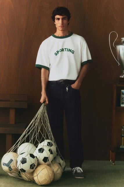 Man posing with a net full of soccer balls in a casual fashion shoot, wearing jeans and a sporty t-shirt in an indoor setting.