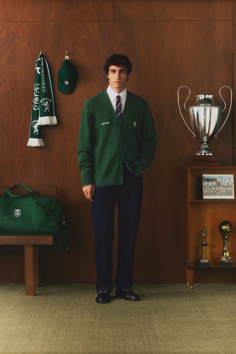 Man in green cardigan stands in front of wooden wall with trophies, a scarf, and a bag, creating a retro fashion and lifestyle scene.