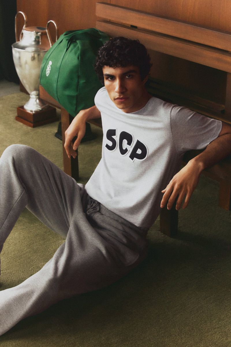 Young man poses while seated indoors wearing a t-shirt and sweatpants, with a wooden bench and trophy in the background.