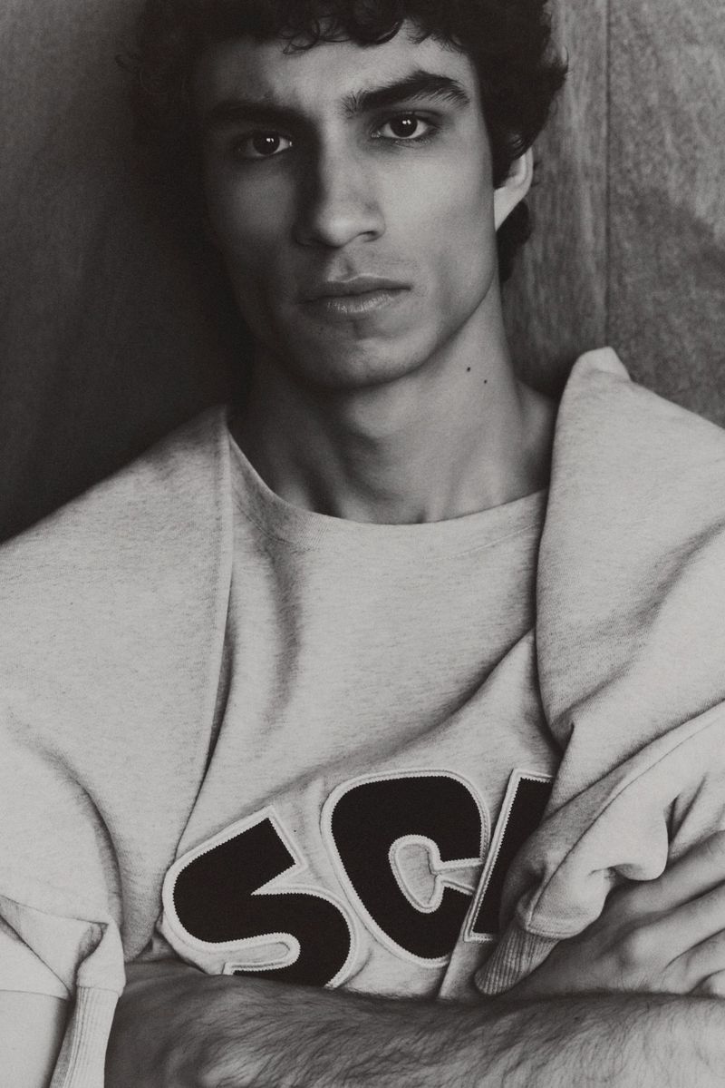 Black and white portrait of an adult male with curly dark hair wearing a sweater with 'SCH' on it, posing indoors against a wall.