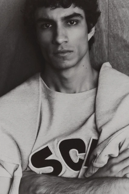 Black and white portrait of an adult male with curly dark hair wearing a sweater with 'SCH' on it, posing indoors against a wall.
