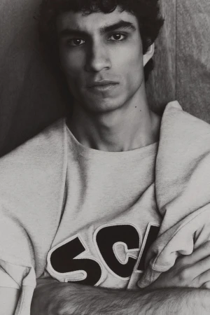 Black and white portrait of an adult male with curly dark hair wearing a sweater with 'SCH' on it, posing indoors against a wall.