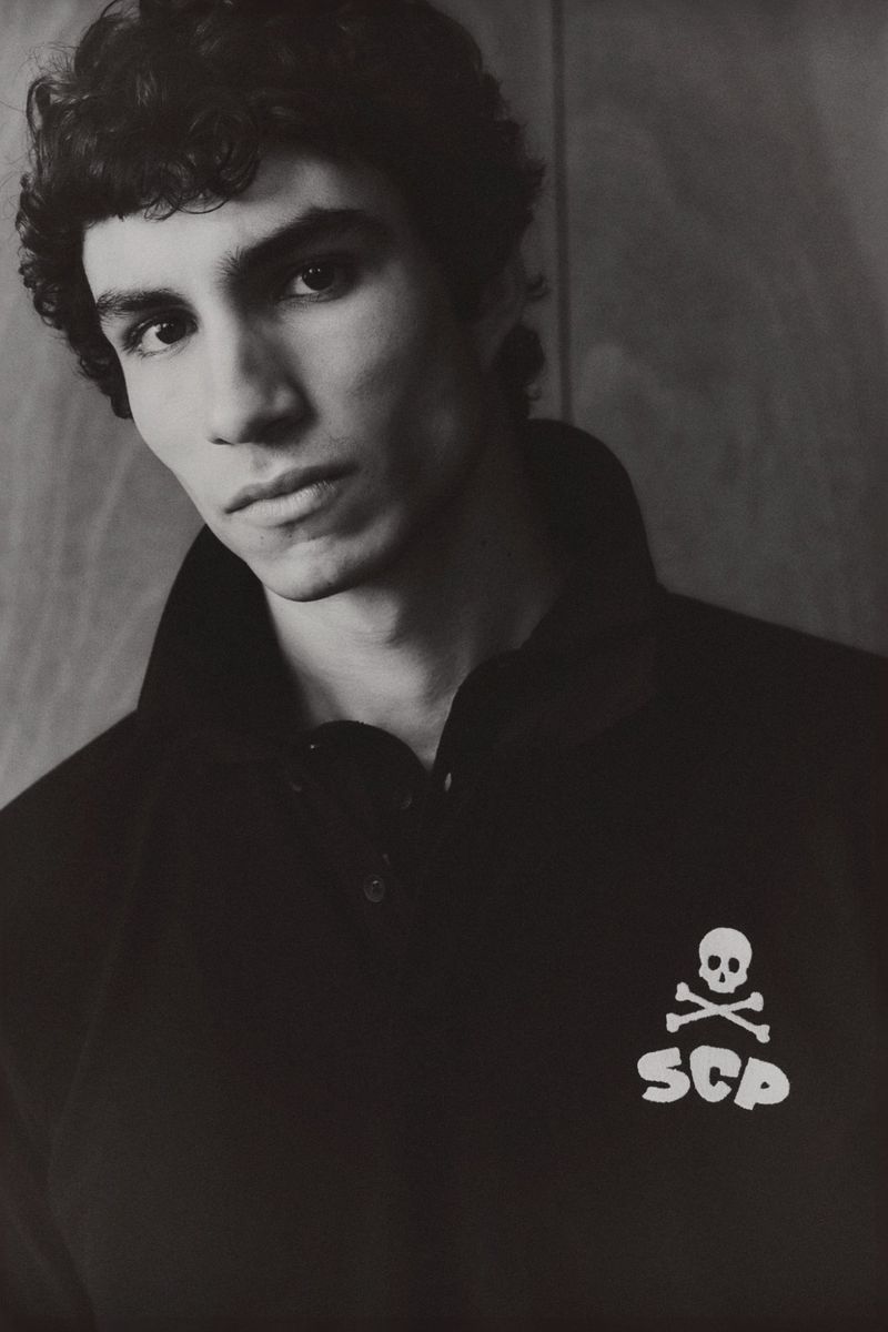 Monochrome portrait of a young man with curly dark hair, wearing a black polo shirt with a skull and crossbones logo.