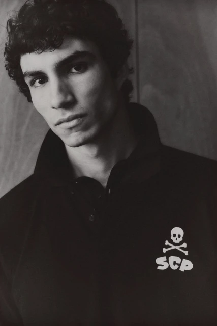Monochrome portrait of a young man with curly dark hair, wearing a black polo shirt with a skull and crossbones logo.