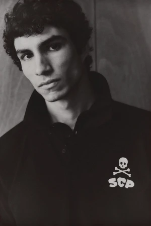 Monochrome portrait of a young man with curly dark hair, wearing a black polo shirt with a skull and crossbones logo.