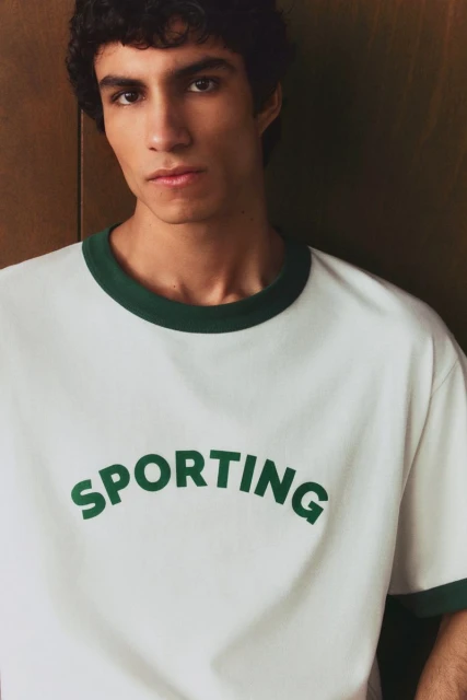 Close-up of a young adult man wearing a white crewneck t-shirt with dark green trim and 'Sporting' printed on the front, indoor setting.