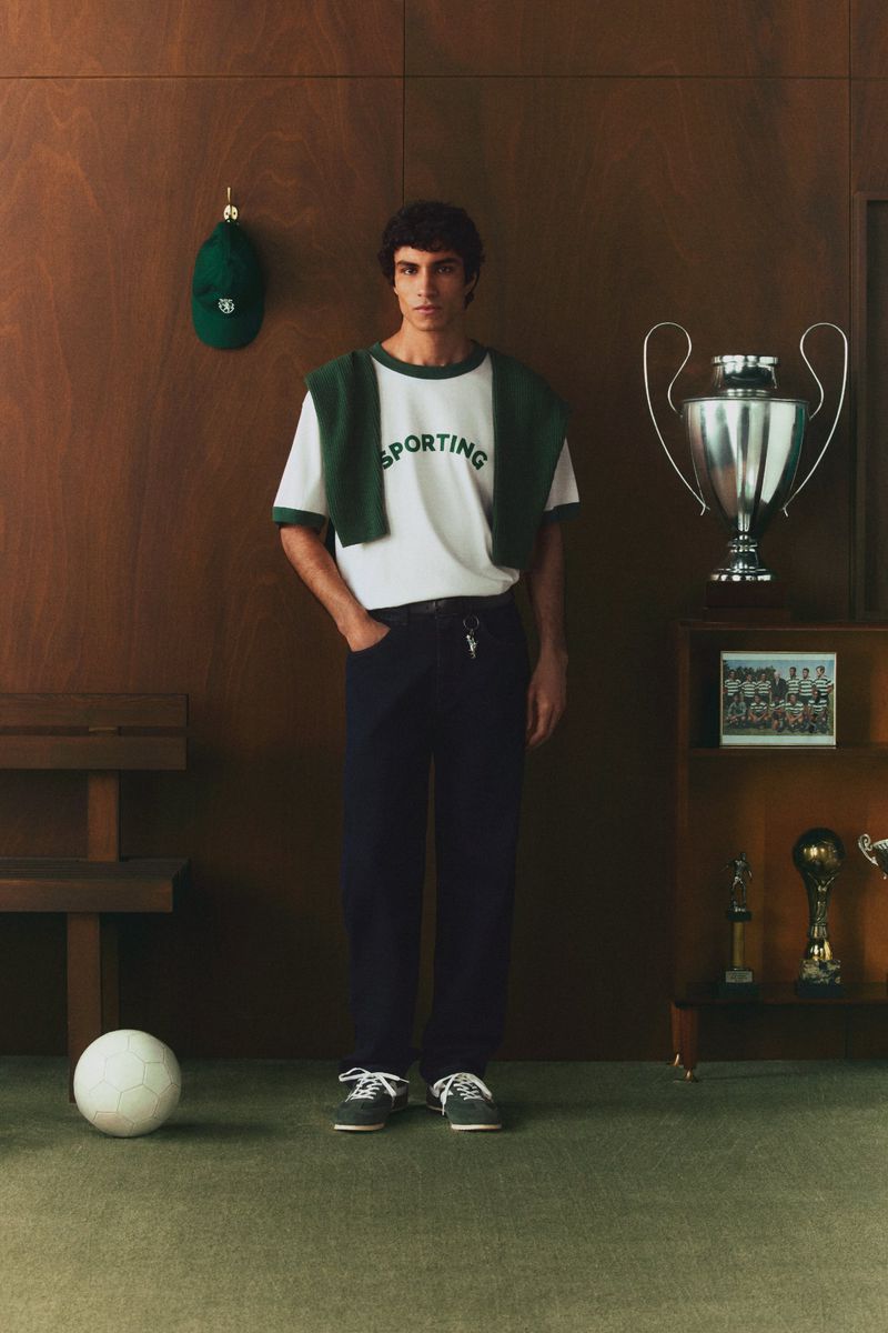 A young man poses indoors wearing a white t-shirt and dark pants, next to a trophy and a soccer ball, with a wood-paneled background.