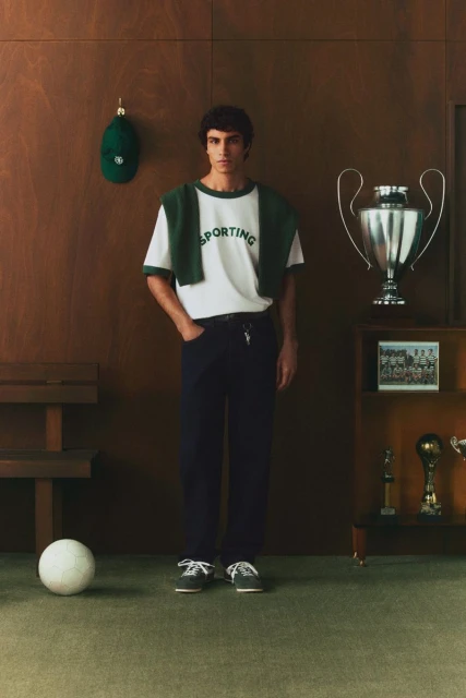 A young man poses indoors wearing a white t-shirt and dark pants, next to a trophy and a soccer ball, with a wood-paneled background.