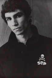 Portrait of a young man with dark curly hair wearing a black jacket with a white skull and crossbones logo.