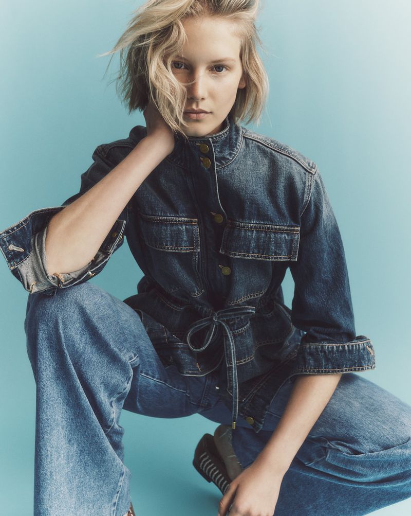 A blonde woman wearing a dark blue denim jacket and matching jeans crouches against a bright cyan studio backdrop.