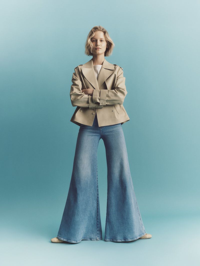 A woman in a trench coat and wide-leg jeans stands with her arms crossed against a graduated blue backdrop, looking at the camera.
