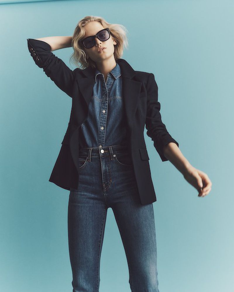 Adult female with sunglasses poses in a denim shirt, black blazer, and blue jeans against a blue backdrop in a studio shot.
