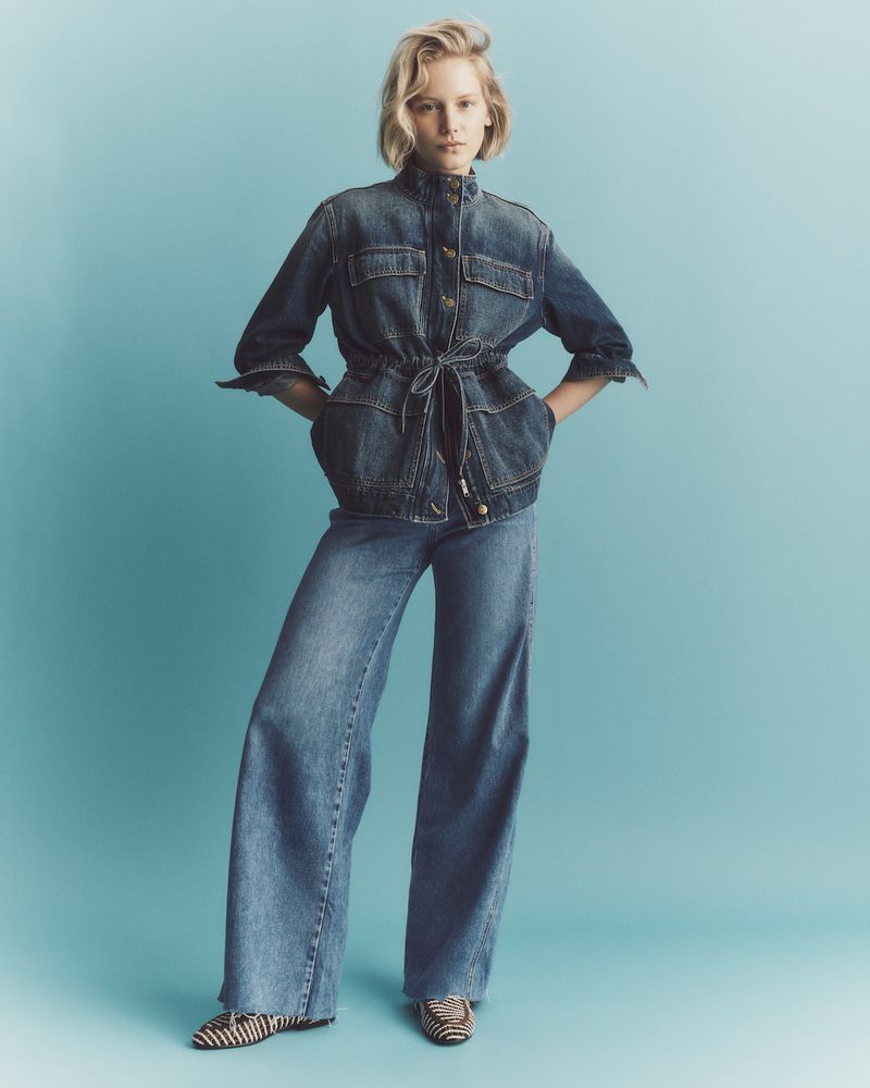 Adult female fashion model poses in a blue denim jacket and wide-legged jeans in a studio shot against a light blue backdrop.