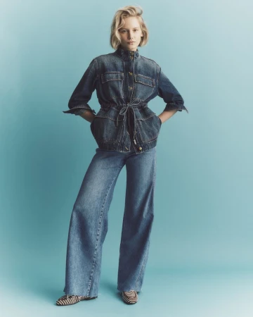 Adult female fashion model poses in a blue denim jacket and wide-legged jeans in a studio shot against a light blue backdrop.