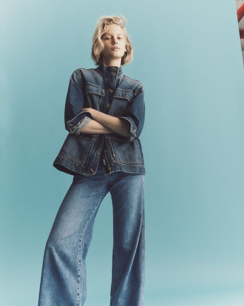 Model with blonde hair wearing a denim jacket and matching jeans against a solid blue background
