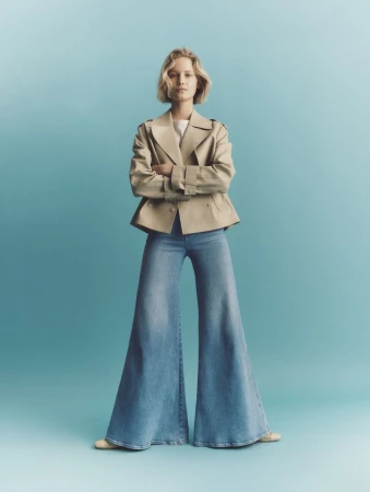 Model standing with arms crossed wearing a cropped beige jacket and wide-leg denim jeans against a soft blue background.