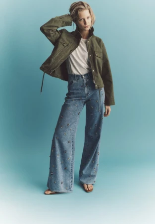 Model in olive green jacket and embellished wide-leg jeans against blue gradient backdrop