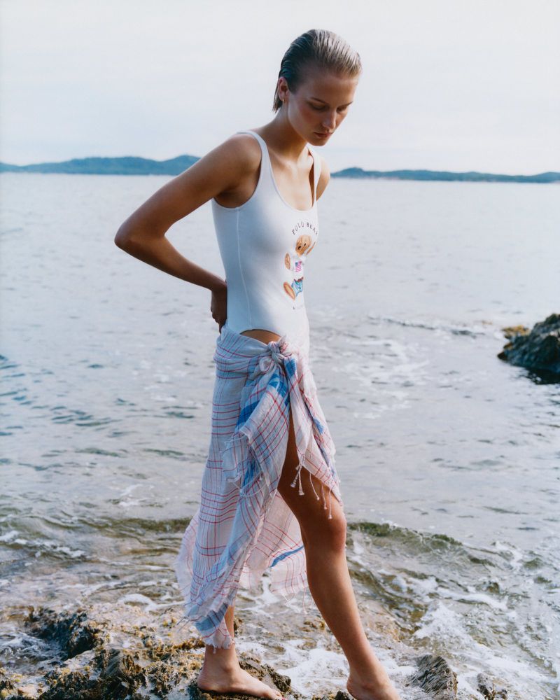 A woman in a white swimsuit and wrap stands on rocks by the sea with the ocean and distant land in the background.