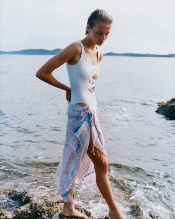 A woman in a white swimsuit and wrap stands on rocks by the sea with the ocean and distant land in the background.