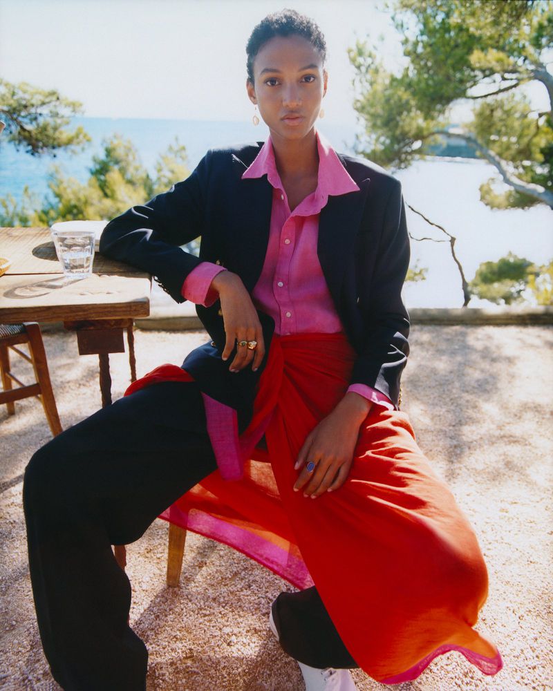 An adult female model wearing a pink shirt, black blazer, and red skirt sits on a chair outdoors by the sea.
