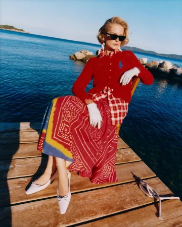 Adult woman in red cardigan, patterned skirt, and sunglasses sits on a dock with water and distant land in the background during daytime.