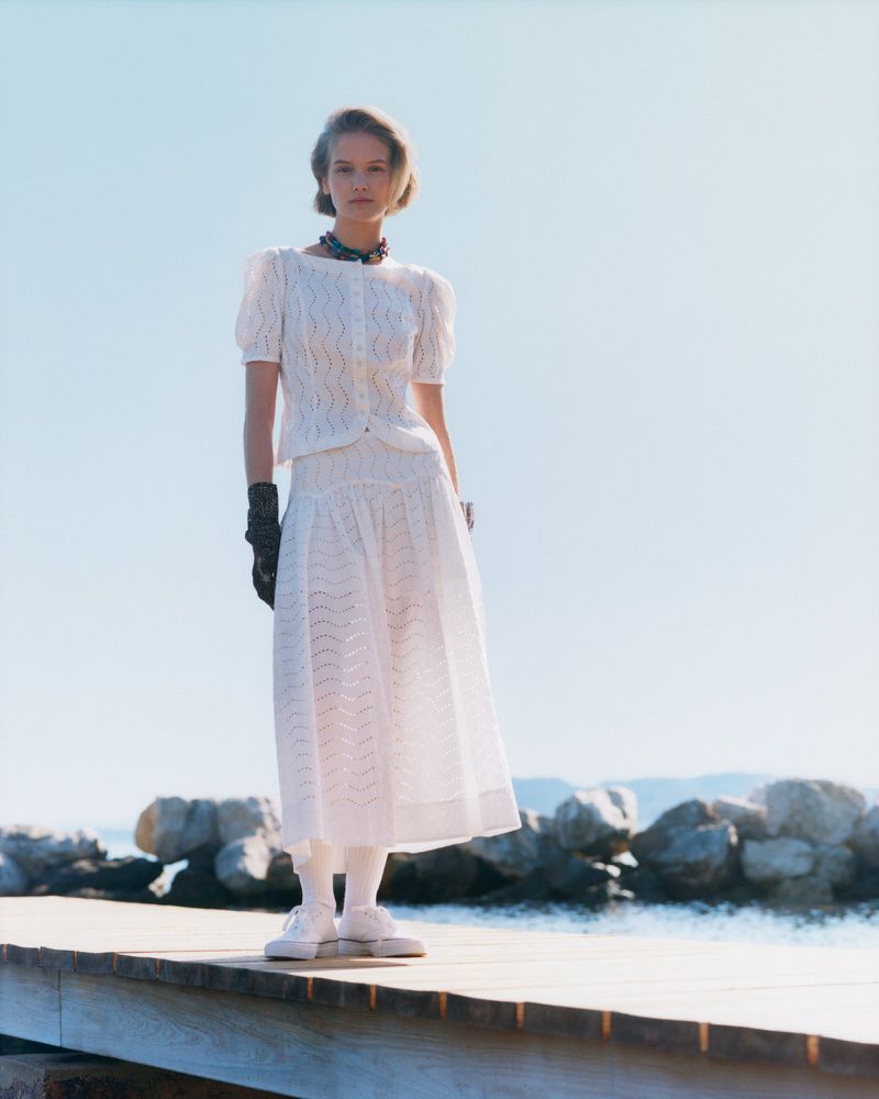 Woman poses on wooden pier wearing white dress, tights, shoes, black gloves, and choker against sky and ocean in fashion shot.