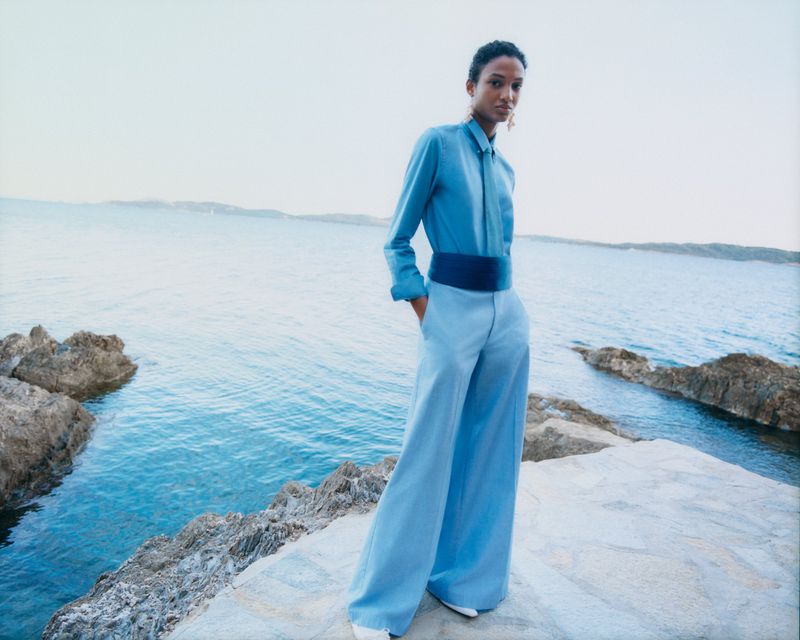 Fashion shot of a woman in blue shirt and pants standing on a rocky coast with ocean view in background, minimalist, natural lighting.