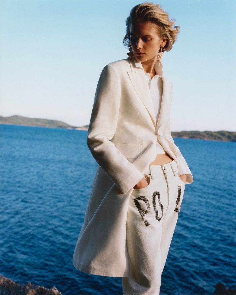 A blonde model wearing a white blazer and white pants with black lettering, standing outdoors by a blue sea at sunset.