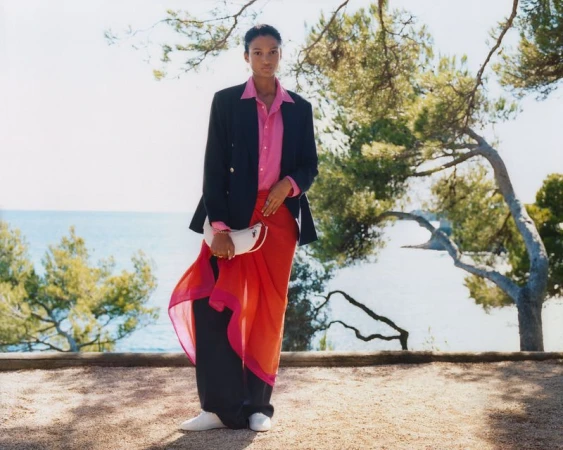 A woman in a pink blouse, dark jacket, and red wrap skirt, standing outdoors on a sunny day, with trees and ocean in the background.