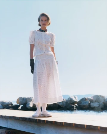 Model wearing a white textured top and matching midi skirt standing on a wooden dock with a clear sky background.