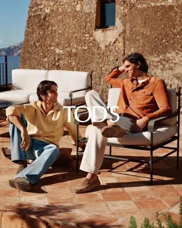 Two adult men are sitting outdoors on neutral furniture with a coastal view in the background, showcasing a relaxed and stylish scene.