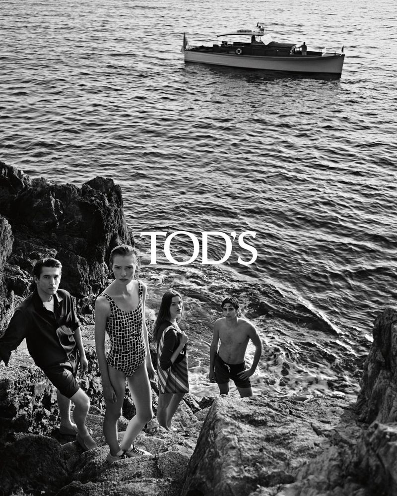 Black and white fashion shot of four people on rocks by the sea with a Tod's logo and a boat in the background.