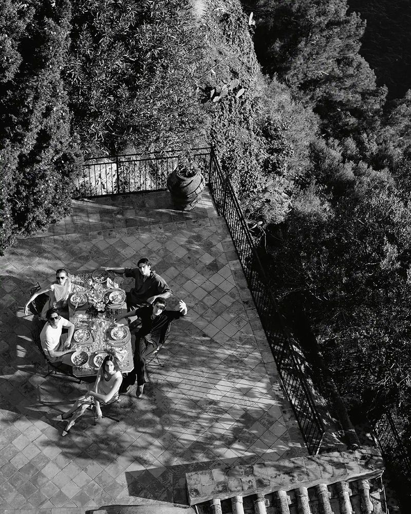 Black and white, high-angle shot showcases a group of six people gathered around a table on a tiled patio with surrounding greenery.