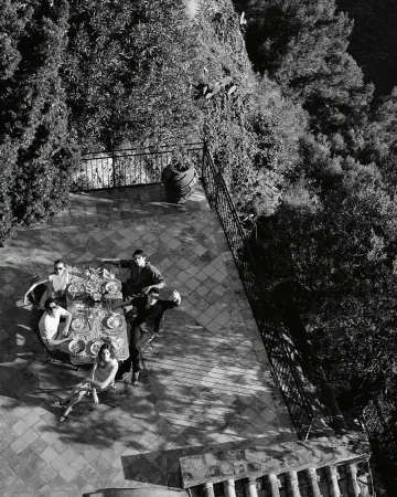 Black and white, high-angle shot showcases a group of six people gathered around a table on a tiled patio with surrounding greenery.