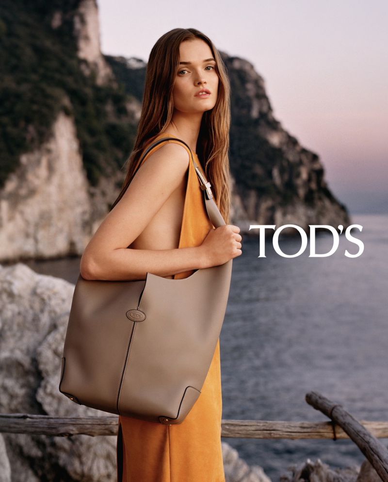 Elegant woman poses with a beige Tod's leather handbag in front of coastal cliffs and ocean, fashion editorial.