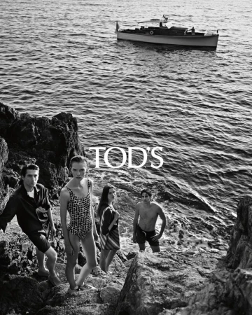 Black and white editorial shot of five models posing near rocky coastal water with a boat visible in the background for Tod's.