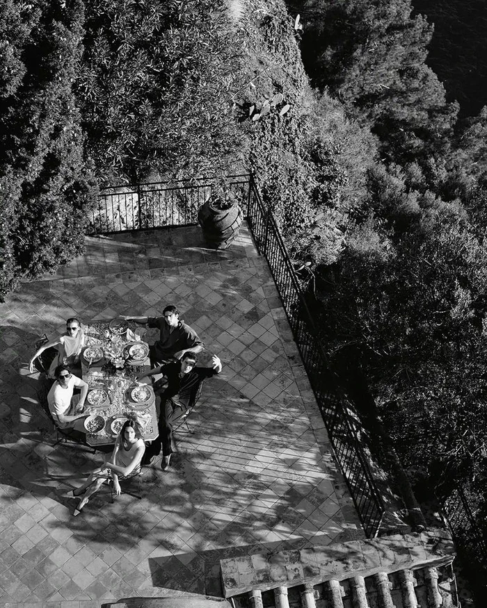 High-angle monochrome photograph of five models gathered around an outdoor dining table on a tiled terrace overlooking dense foliage.
