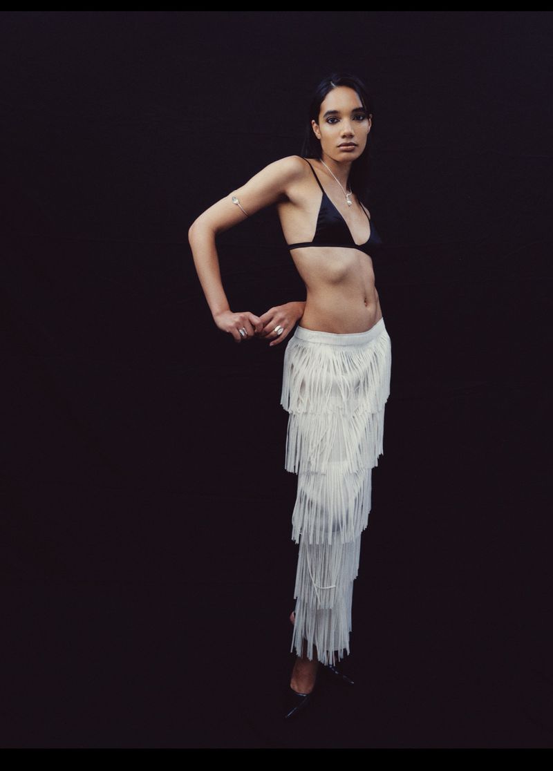 Woman in black bikini top and fringed skirt poses against dark background in studio. Minimalist fashion shot with a modern aesthetic.