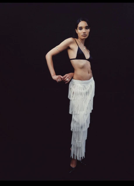 Woman in black bikini top and fringed skirt poses against dark background in studio. Minimalist fashion shot with a modern aesthetic.