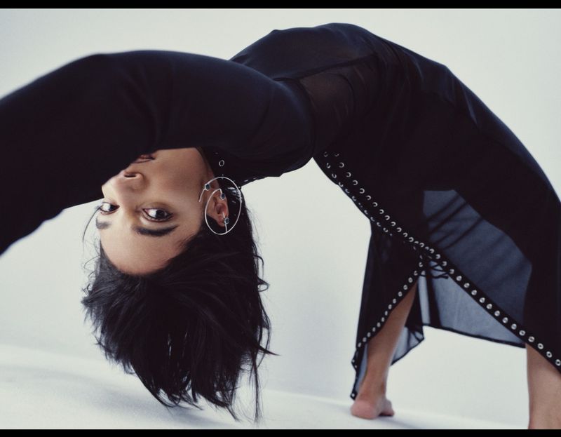 Adult female arches backward wearing a long black sheer dress in a studio. The model's face is visible, with dark hair and silver earrings.