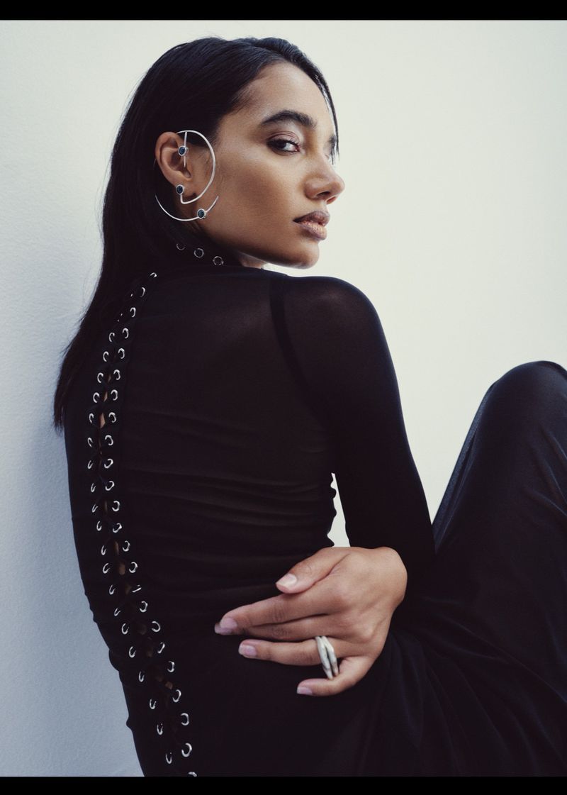 Portrait of a young woman with dark hair wearing a black top with lace-up detail, posing against a neutral wall in an indoor setting.