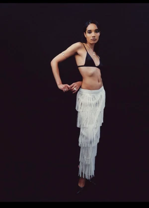 Model in black bralette and white fringed skirt standing against a dark background for the CD Jewelry Campaign.