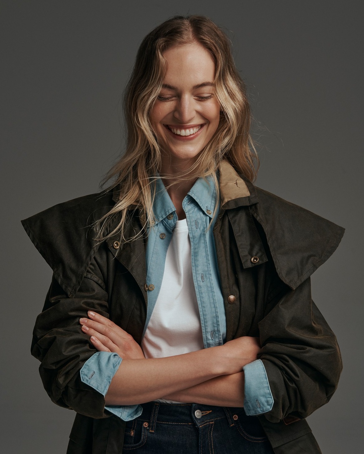 Smiling blonde woman in a studio portrait wearing a green hooded jacket, light blue shirt, and dark jeans with crossed arms.