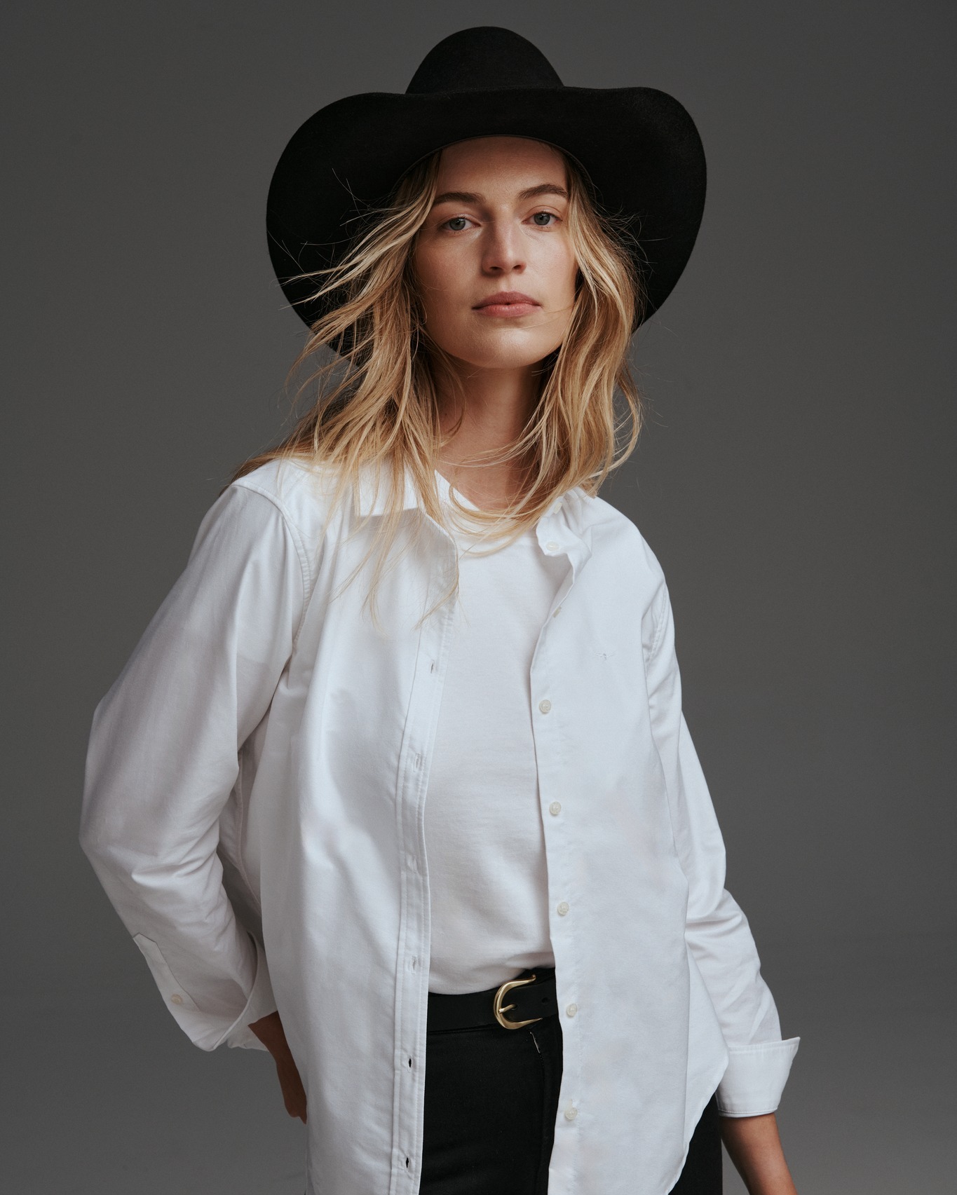 A blonde woman poses in a white button-down shirt and black hat against a gray background, exuding a stylish and confident look.