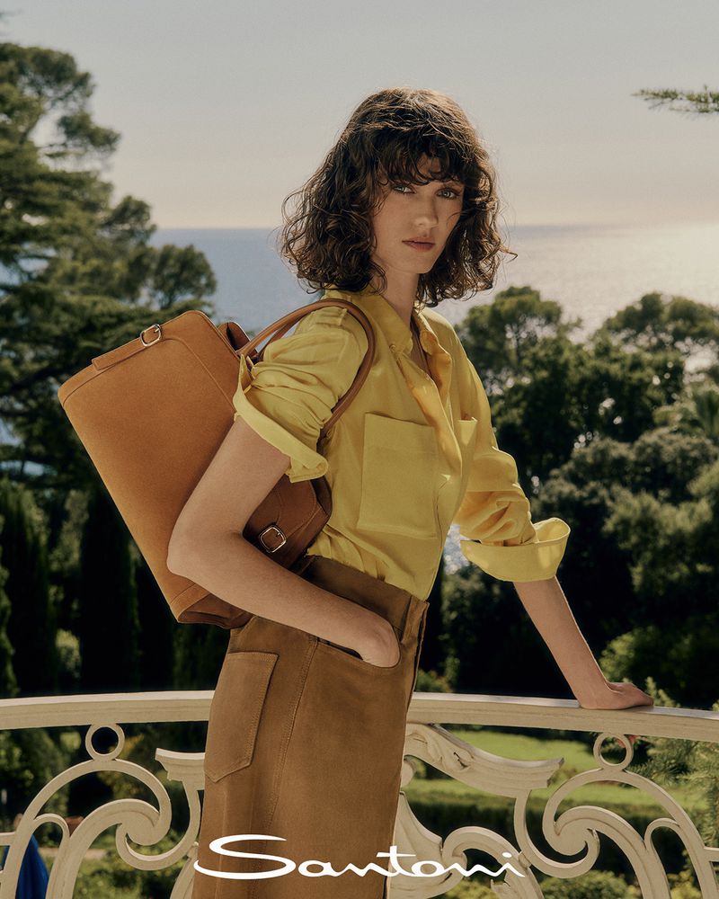 Brunette woman in yellow shirt and brown skirt poses with leather bag on balcony with an ocean view, outdoor fashion photoshoot.