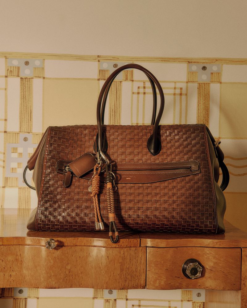 Brown woven leather tote bag with brown handles, leather tassels and tags hanging on a wooden table against a patterned wallpaper.